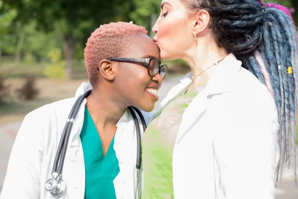 Пара лікарів. Дві красиві дівчата, біла дівчина з дредлоками, цілують чорну дівчину в лобі. У білих халатах з фонендоскопами. На відкритому повітрі — стокове фото