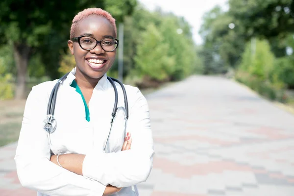 Портрет молодої чорної дівчини, лікаря в білій шубці, з фонендоскопом. Посміхаючись дивитися на камеру, на відкритому повітрі — стокове фото