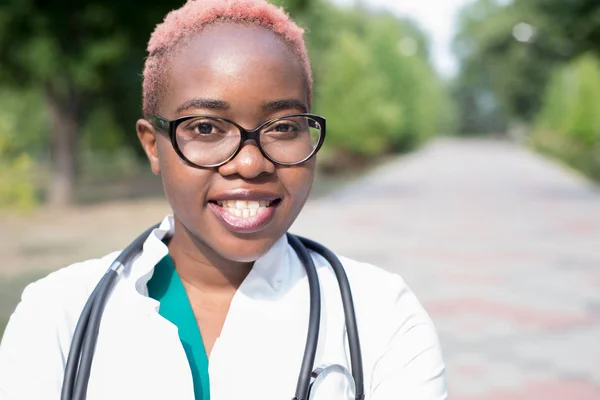 Портрет молодої чорної дівчини, лікаря в білій шубці, з фонендоскопом. Посміхаючись дивитися на камеру, на відкритому повітрі — стокове фото