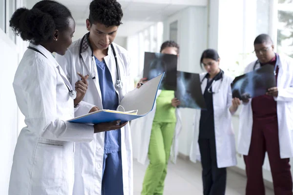 Een groep van gemengde ras artsen. Jongeren in witte jassen, die in de ziekenhuisgang staan, glimlachen en houden medische documenten in hun handen. Bespreek medische gezondheidskwesties — Stockfoto