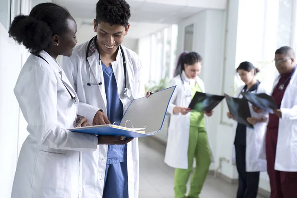 Група лікарів змішаної раси. Молоді люди в білих халатах, стоячи в лікарняному коридорі, посміхаються, тримаючи в руках медичні документи. Обговорити питання медичного здоров'я — стокове фото