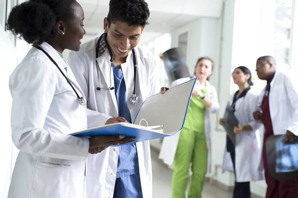 Bir çift doktor, melez ırk. Beyaz önlüklü, doktor, gülümseyen genç insanlar ellerinde tıbbi belgeler tutuyorlar. Arka planda bir grup doktor röntgenleri inceliyor.