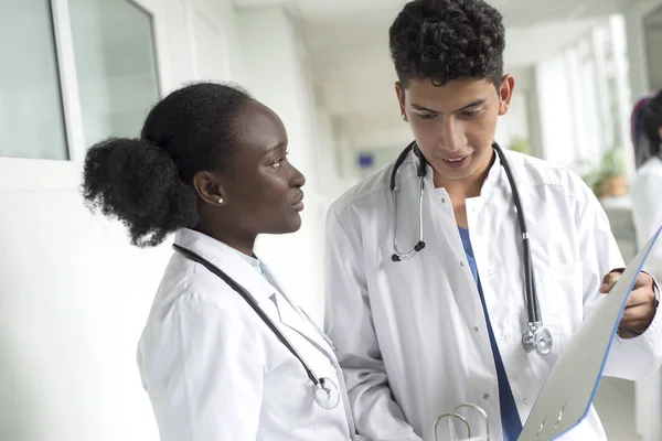 Black woman and white male doctors. A pair of young doctors of mixed race in white coats with a folder in their hands discussing medical issues. In the corridor of the hospital — 스톡 사진