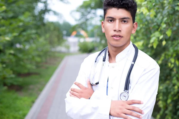 Портрет белого врача. Эквадорский парень в белом халате с фонендоскопом улыбается. На открытом воздухе — стоковое фото