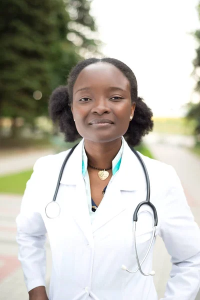 Ritratto di una graziosa ragazza afroamericana. Medico in cappotto bianco, con un fonendoscopio, sorriso, per strada — Foto Stock