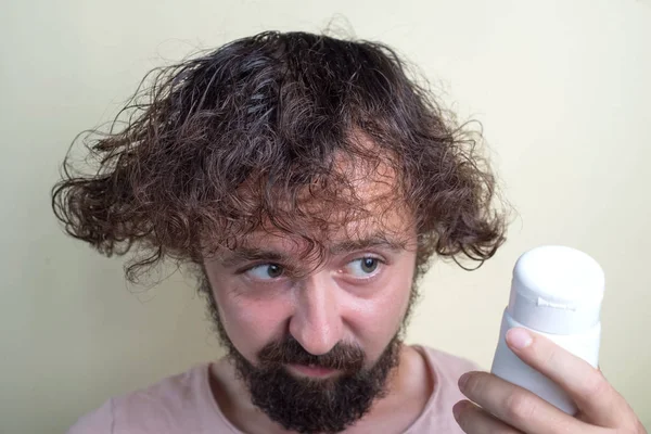 Portrait of a young guy with long dirty hair and problematic scalp. With disbelief looks at a bottle of shampoo.