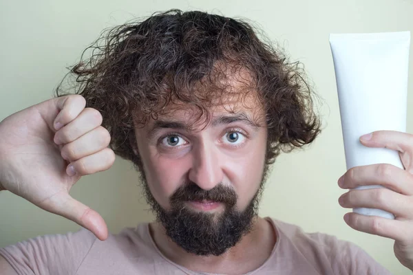 Ritratto di un ragazzo con capelli sporchi e cuoio capelluto problematico. Tenendo in mano un cattivo shampoo, mostrando il pollice verso il basso — Foto Stock