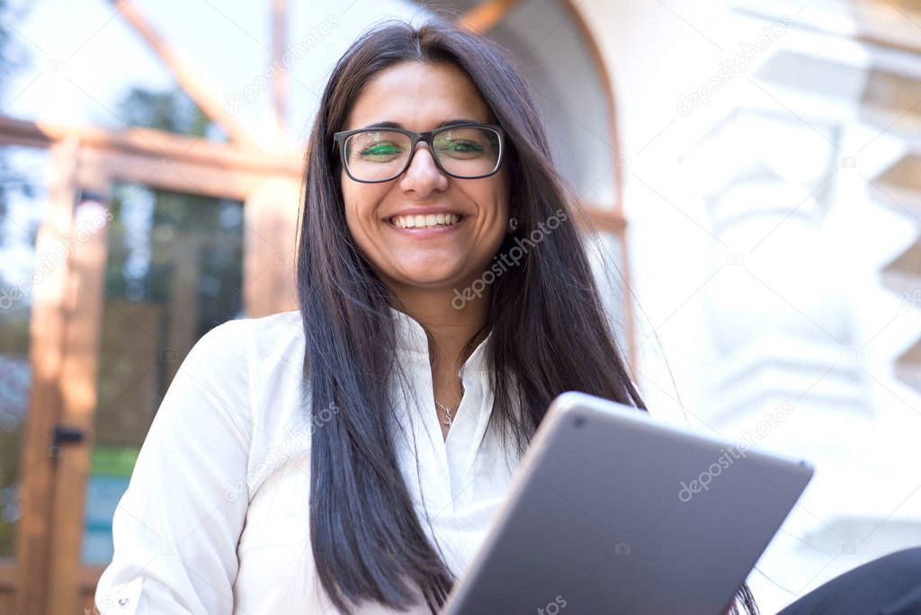 portrait of a beautiful indian girl. Business woman, wearing glasses, smiling, sitting on the steps of an office building, holding a tablet in her hand