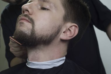 Berber dükkanında oturan sakallı bir gencin yakın plan portresi. Usta sakal stili yapıyor.