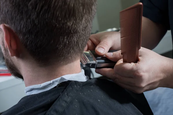 Frisören klipper hår med en maskin för en ung kille med skägg. Närbild av en mästare hand skära en mans hår — Stockfoto