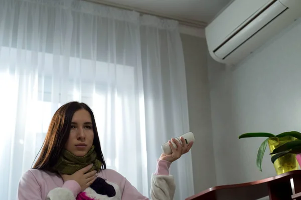 Ev yapımı pijama giyen, boğazı ağrıyan, öksüren klima arka planında güzel bir kızın portresi. Uzaktan kumandayı elimde tutuyorum, kapatmaya çalışıyorum.