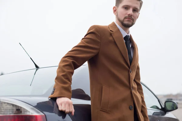 Close-up portret van een bebaarde jongeman in een zakelijk stijlvol pak en met een stropdas. Een zakenman staat bij de auto.. — Stockfoto
