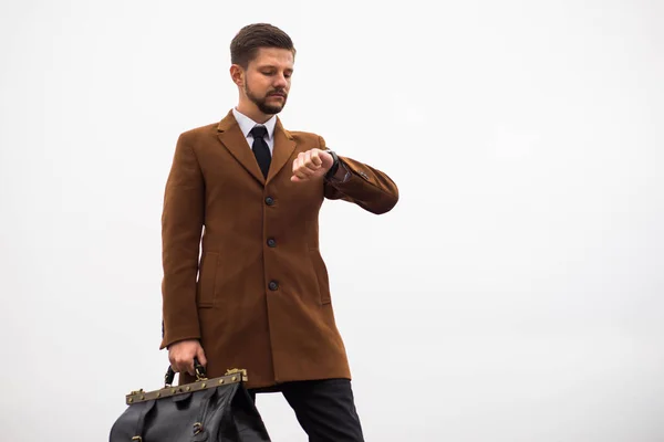 Portret van een jongeman van dertig, in een zakelijke stijl, met een leren tas in zijn handen, kijkend naar zijn horloge. Tegen de lucht, straatstijl — Stockfoto