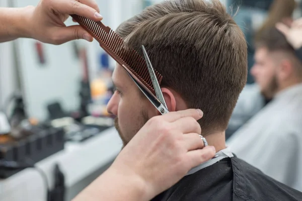 Mäster frisör klipper hår på huvudet på en ung skäggig kille. Närbild av handen på en professionell hårklippare med sax i en frisör butik. — Stockfoto