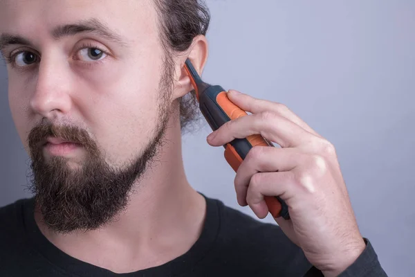 Retrato de um jovem barbudo a usar um aparador para cortar cabelo nas orelhas. Em um fundo cinza — Fotografia de Stock