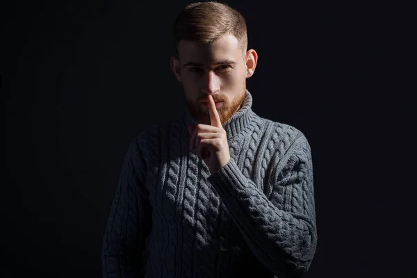 Portret van een jonge man met baard van vijfentwintig jaar. In een warme wintertrui, trots, grijs, kijkend naar de camera en met een vinger naar zijn mond, met de achtergrond voor een roep om stilte. — Stockfoto