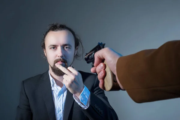 Närbild porträtt av en man som visar sitt långfinger, vars ansikte pekar sin hand med en annan mans pistol. På en grå bakgrund. Begreppet dödande. Dramatisk bild. — Stockfoto