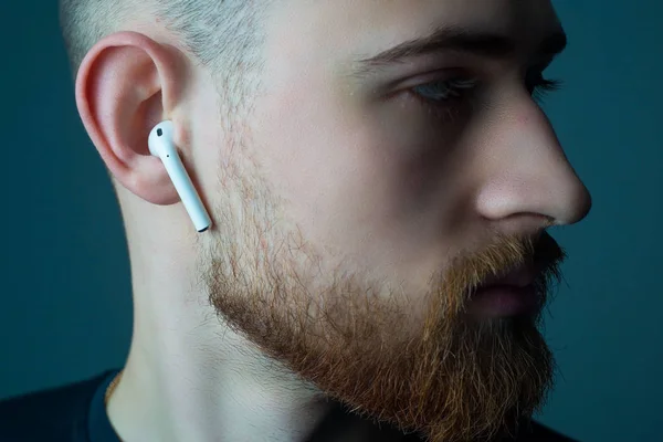 Close-up portrait of a young bearded guy of twenty-five years old, looking at the camera, in wireless white headphones in his ears. — Stock Photo, Image