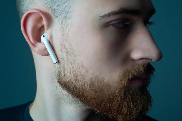 Close-up portret van een jonge man met een baard van vijfentwintig jaar oud, kijkend naar de camera, in draadloze witte koptelefoon in zijn oren. — Stockfoto