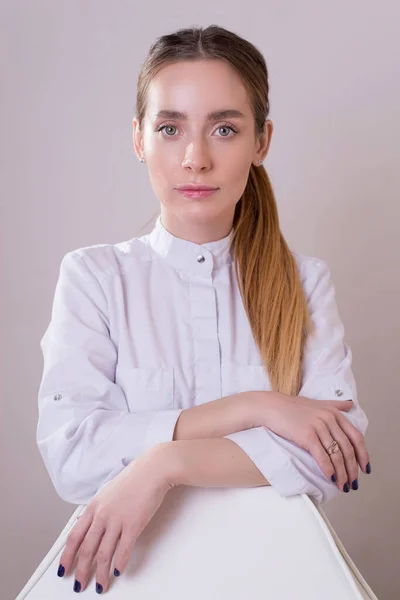 Retrato de un médico cosmetólogo en un consultorio detrás de un lugar de trabajo. Hermosa chica con ojos expresivos en un abrigo blanco mira a la cámara —  Fotos de Stock