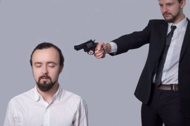 Takım elbiseli bir iş adamı beyaz gömlekli başka bir adamın kafasına silah doğrultuyor. Gri bir arka planda. Öldürme kavramı.