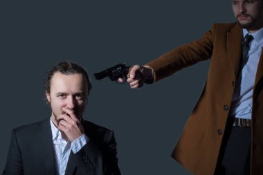 Takım elbiseli bir iş adamı beyaz gömlekli başka bir adamın kafasına silah doğrultuyor. Gri bir arka planda. Öldürme kavramı.