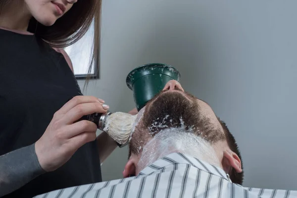 Närbild av handen på en flicka till en professionell mästare i skägg, en rakborste och tvål kommer att skumma skägget av en ung man, förbereda henne för rakning — Stockfoto