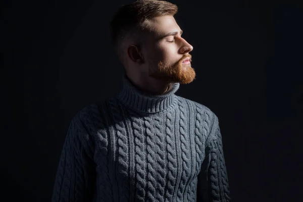 Studio portret van een jonge man met baard van vijfentwintig jaar oud. In een warme wintertrui onder een trotse, grijze kleur, sluit zijn ogen, draait zijn hoofd naar de zijkant. Op een zwarte achtergrond. — Stockfoto