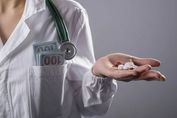 Close-up of a doctor in a white coat, with a phonendoscope, in his pocket a press of dollar bills, in his hand medical pills. On a gray background, studio photo. Bribe concept