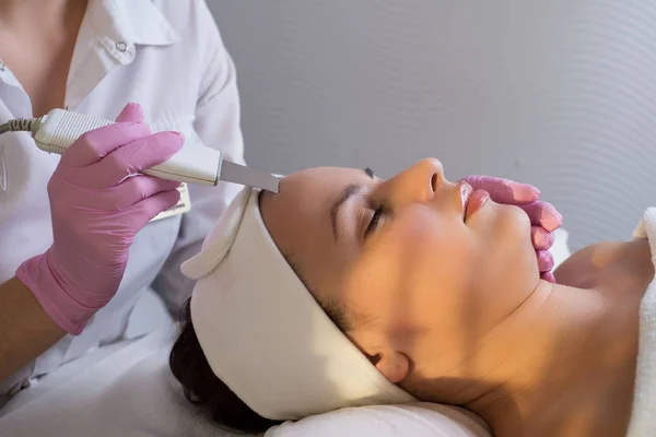 Close-up, esteticista profissional usando um limpador facial ultra-sônico, limpa a pele de um paciente menina, em um salão de beleza — Fotografia de Stock