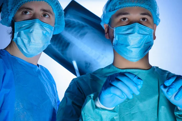 Retrato de dos cirujanos con batas estériles, sombreros y máscaras. Un cirujano, preparándose para operar, sostiene un bisturí con un soporte de aguja en las manos, otro médico sostiene una radiografía del paciente — Foto de Stock