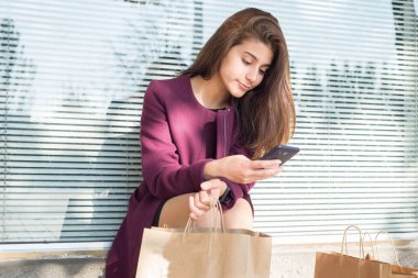 Pencerenin yanında güzel bir Hintli kız oturuyor. Elinde akıllı bir telefon ve yanında çantalar var. Perdelerin arkasında.