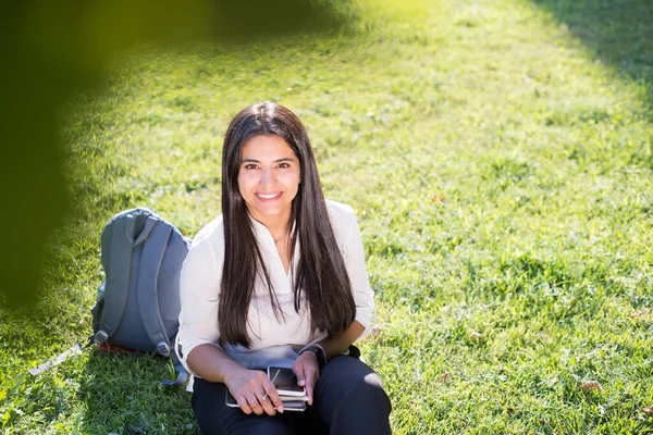 Indian girl, student, sitting on the green grass, with a backpack, smiling and holding a tablet