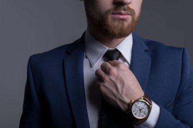 İş elbisesi içinde genç sakallı yakışıklı bir adamın portresi, kameraya bakıyor, kol saatini gösteriyor, deri bir kayışla. Reklamcı erkek saatleri. Gri bir arka planda. İş adamı portresi