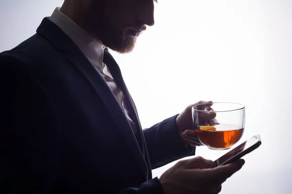 close-up, on a clearance on a white background, the silhouette of a bearded handsome guy drinking tea from a transparent cup, uses a smartphone. Promotional photo of black tea. Coffee break man. Lunch. Tea party Bearded guy is drinking tea on a white