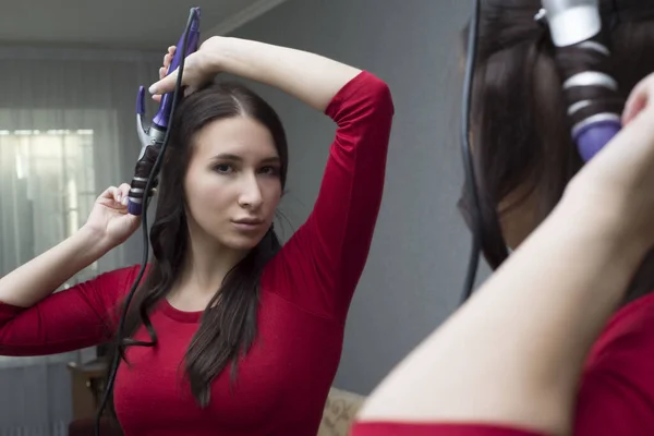 portrait of a beautiful young girl, with flawless hair, stands at home by the mirror, twists her hair with a curling iron. On a gray background. Woman in red dress curls hair with curling iron