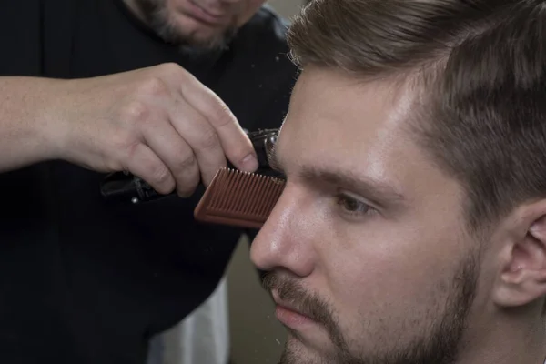 Hairdresser cuts hair with a trimmer for a young guy with a beard. Close-up of a master haircutting male hair. Cutting hair at the temples of a guy. Bearded man at the hairdresser. Men\'s haircut