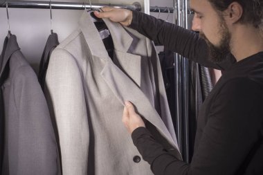 Bin yıllık genç sakallı bir adamın erkek giyim mağazasındaki yakın plan portresi iş için bir takım elbise seçiyor. Erkek giysisi satışı. İş ceketi. Gri ve bej renkli takım elbise. Bir adam takım elbisesinin kalitesini ölçüyor.