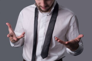 A portrait of a young bearded man in a white shirt indignantly shrugs, pointing to a tie, amid the inability to tie a tie correctly. On a dark gray background. Business dress code, a man is going to an important meeting. Demonstration of masculinity,
