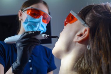 Profesyonel diş beyazlatma prosedürünü uygulayan bir dişçi doktoru portresi ultraviyole radyasyon kullanıyor. Koruyucu turuncu gözlüklü hasta ve doktor. Gri bir arka planda. Dişçi kliniğinde. Hastalıkların önlenmesi ve tedavisi