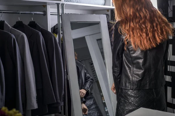 portrait of a beautiful red-haired woman thirty-seven years old, in a women\'s clothing store, a girl trying on a leather jacket made of high-quality fabric by the mirror. Sale of women\'s clothing in a boutique. Shopping, buying new things, shopaholic