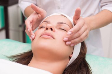 Kız masaj terapisti güzel bir kadınla yüz masajı yapıyor. Eller kapanıyor. Masaj odası. Spa yüz bakımı. Yüzün kasları için egzersizler. Rahatlatıcı yüz masajı