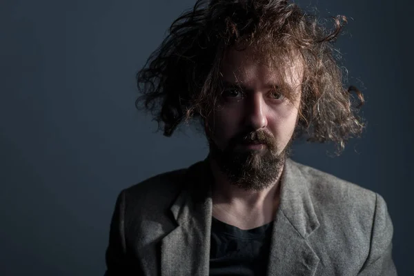 Studio dramatic portrait of a young bearded guy, thirty years old, with curly dirty greasy hair and a jacket. On a gray background. The concept of not washed hair. Dirty hair. Dramatic light. Promotional photo of a man with long hair. Hair and scalp