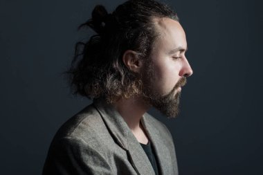 30 yaşında, kıvırcık, yağlı saçlı ve ceketli, sakallı bir gencin dramatik bir portresi. Gri bir arka planda. Saç yıkamama kavramı. Kirli saçlar. Dramatik ışık. Uzun saçlı bir adamın promosyon fotoğrafı. Saç ve kafa derisi. 