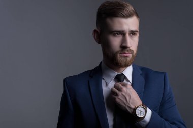 İş elbisesi içinde genç sakallı yakışıklı bir adamın portresi, kameraya bakıyor, kol saatini gösteriyor, deri bir kayışla. Reklamcı erkek saatleri. Gri bir arka planda. İş adamı portresi