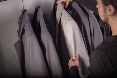 Bin yıllık genç sakallı bir adamın erkek giyim mağazasındaki yakın plan portresi iş için bir takım elbise seçiyor. Erkek giysisi satışı. İş ceketi. Gri ve bej renkli takım elbise. Bir adam takım elbisesinin kalitesini ölçüyor.