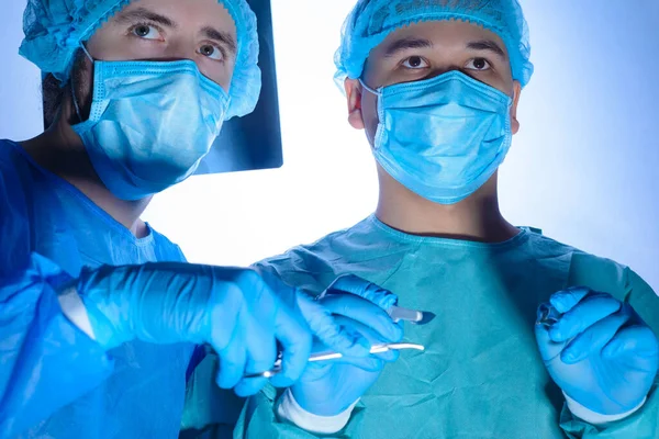 Portrait of two surgeons in sterile gowns, hats and masks. One surgeon, preparing to operate, holds a scalpel with a needle holder in his hands, another doctor assisting the first surgeon. Against the background of the operating lamp. Studio photo me