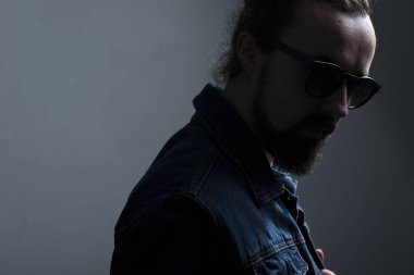 Gözlüklü, kot ceketli, gri arka planda sakallı genç bir adamın dramatik gölge portresi. Dramatik ışık. Esmer adam portresi. Güneş gözlüklü bir adamın promosyon fotoğrafı.