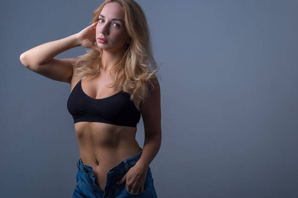 studio portrait of a young beautiful blonde girl, twenty-eight years old. In jeans and a black bra top. Advertising photo on a gray background. Free space. Jeans style for girls. Beautiful blonde topless, bra, women's sports topic.