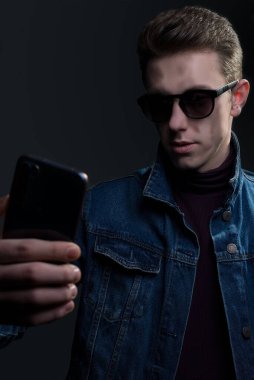 Closeup portrait of a young guy, in sunglasses, holding a smartphone, taking a selfie. Denim style. on a gray background.